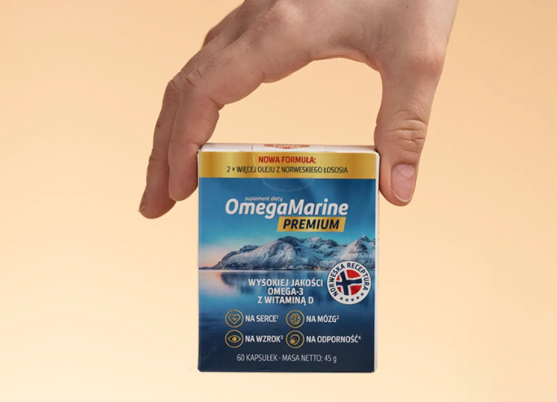 Natural Pharmaceuticals OmegaMarine™ Premium Dłoń trzyma pudełko suplementu diety OmegaMarine™ Premium — wysokojakościowe omega‑3 z witaminą D; nowa formuła: 2× więcej oleju z norweskiego łososia.