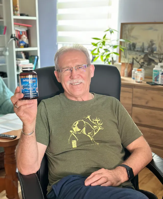 Mężczyzna siedzi w fotelu i trzyma butelkę suplementu OmegaMarine™ Premium; domowe wnętrze w tle.